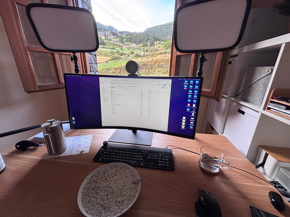 A curved ultrawide monitor on a wooden desk, flanked by two video lights, with a bowl of porridge in the foreground and an open window behind showing a green Andorran valley