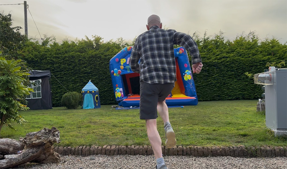Niall from behind in a checked shirt and shorts, mid-stride across a lawn towards an inflatable bouncy castle, with a hedge and a small play tent either side