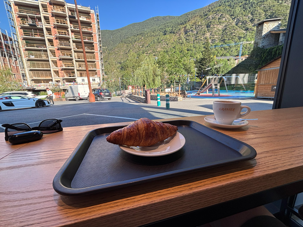 A croissant on a tray and a cup of coffee on a cafe window counter, looking out at a playground, an apartment block under construction and a forested mountainside