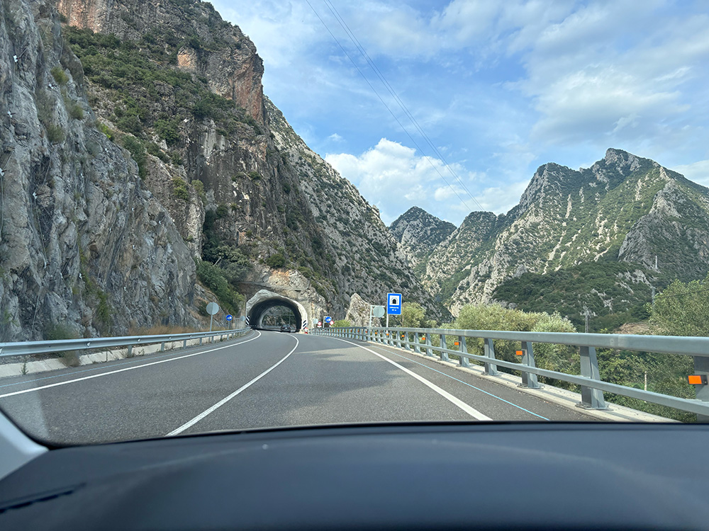 The view through a car windscreen of a motorway curving towards a tunnel mouth, cut into steep limestone crags under a blue sky