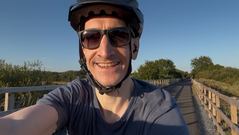 Niall on the Slieverue Greenway in helmet and sunglasses, the fenced path and low sun running off behind him