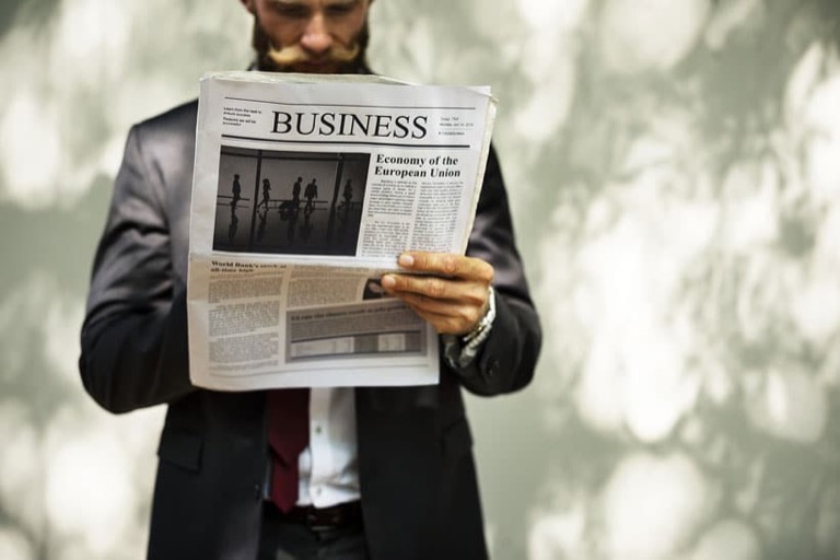 A man in a suit holding up the business section of a newspaper, his face hidden behind it