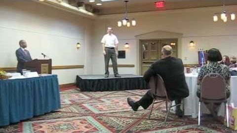 Niall on a low stage in a hotel function room, the contest chair at a lectern to one side and the audience at round tables