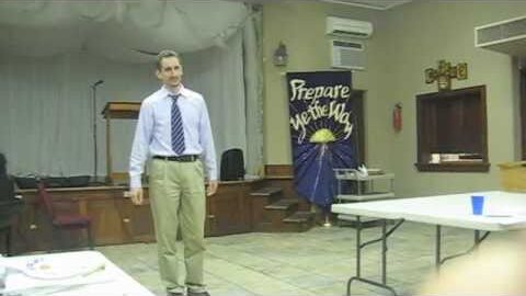 Niall in shirt and tie standing in front of a church hall stage, a Prepare Ye The Way banner behind him